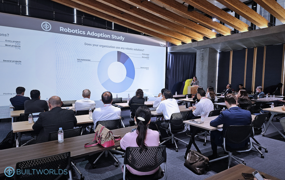 A woman stands behind a podium speaking to roughly twenty business professionals, with a powerpoint showing a pie chart projected on a large screen. 