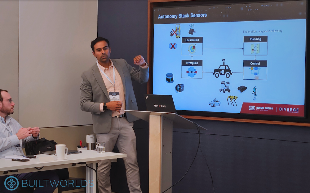 A man with dark hair in a gray suit raises his hand as he presents in front of a large screen showing a presentation. 