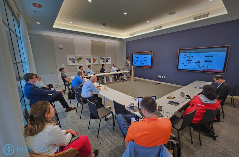 A man in a suit stands in front of an audience of business professionals in a modern conference room presenting on equipment and robotics.