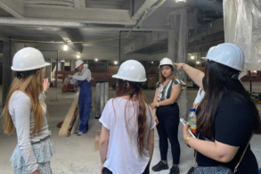 Women wearing white hardhats visit an active jobsite for the construction of a building.