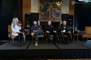 A woman in white speaks into a microphone while three men sit to her right, ready to answer questions.