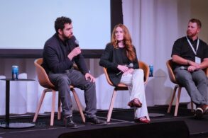 A man leans onto his knee as he speaks into a microphone while two other speakers look on.
