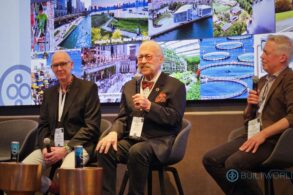 A man with a large handlebar mustache talks into a microphone as two men on either side listen.