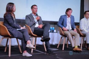 A panel of speakers sits in orange armchairs as a man speaks into a microphone