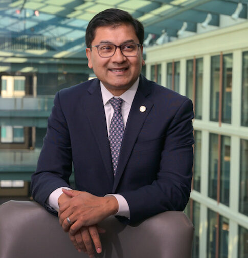 A man in a blue suit with a pin on the lapel wearing glasses smiles looking off camera.