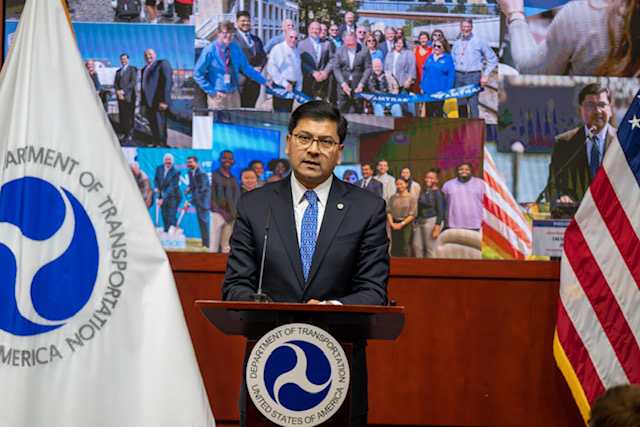 A man in a dark suit talks at a podium flanked on either side by the American flag and flag of the US Department of Transportation.