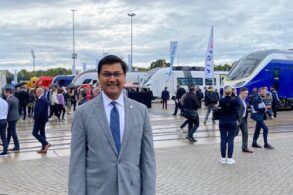 A man in a gray suit smiles in front of several large, colorful, modern trains.