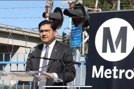 A man in a dark suit speaks into a microphone in front of a Metro sign.