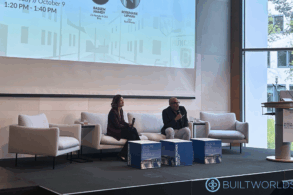 Two people smile and speak to each other while seated on a couch on a platform stage.
