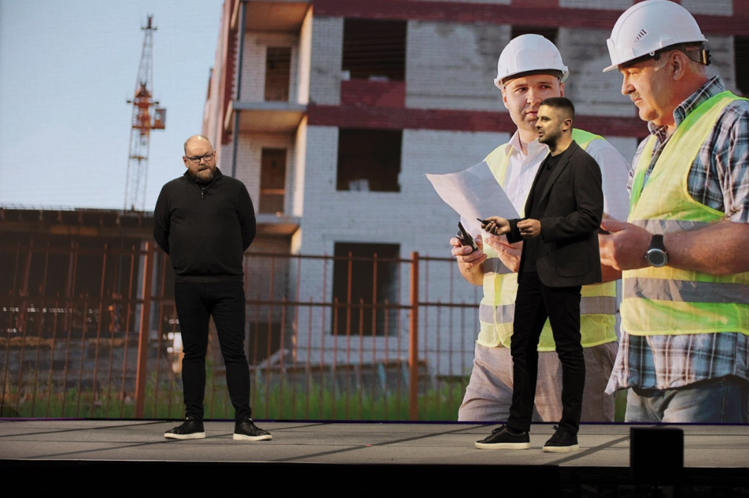 two professionals giving a presentation on stage in front of a large screen showing construction professionals reviewing plans